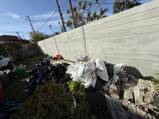 Bagged garden waste, filled white demolition bags and broken concrete rubble lined up along a poured concrete wall in a Los Angeles County back yard