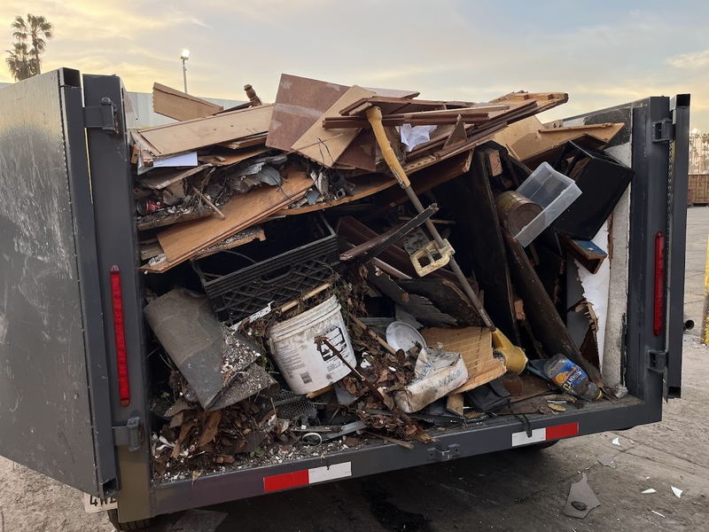 Our dump trailer loaded with mixed construction debris at dusk: cut timber, baseboard, a paint bucket, crates and broken fixtures stacked above the tailgate on a Los Angeles County job