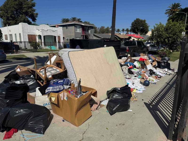 A large curbside pile on a Los Angeles County street: a stained mattress on its side, dining chairs, collapsed cartons, black bags and loose trash spread across the sidewalk in front of the houses