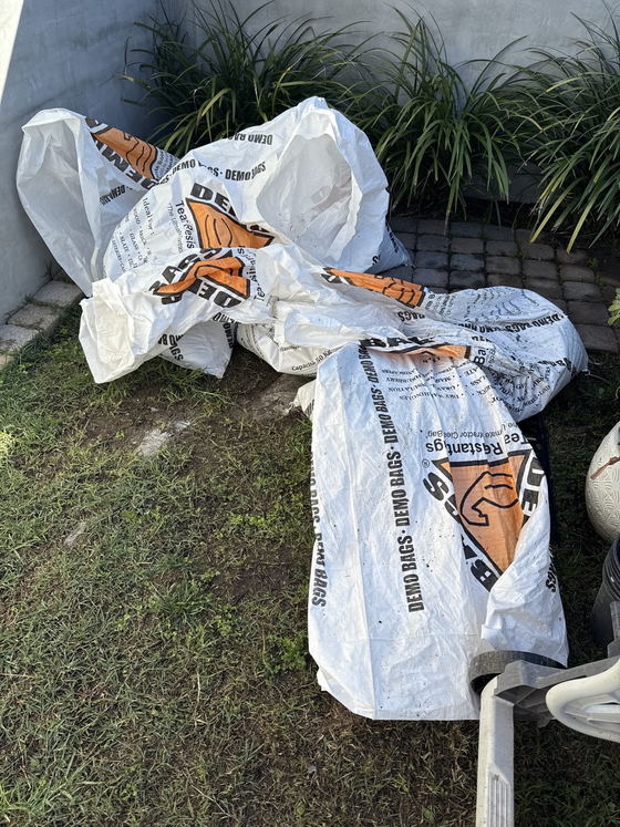 Heavy-duty white demolition bags packed full of renovation waste lying on the lawn and paved patio of a Los Angeles County property, staged for a hand carry to the trailer