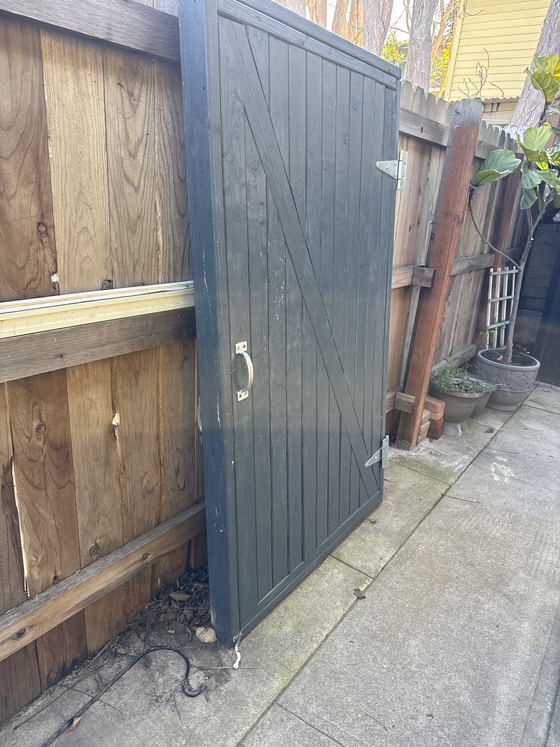 A heavy timber garden gate lifted off its hinges and leaned against a failing board fence along a narrow Los Angeles County side path, ready to be carried out