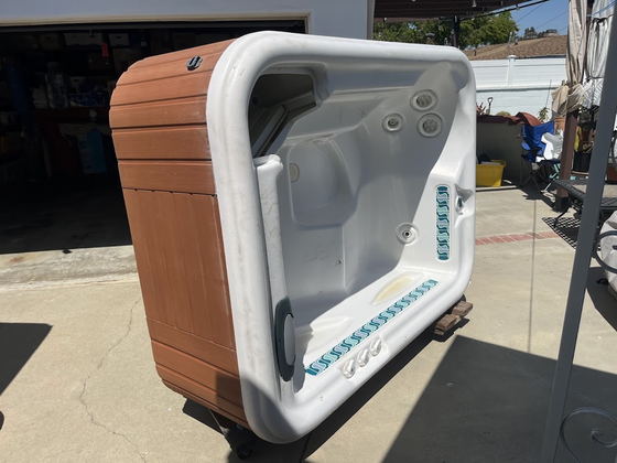 A four-seat hot tub tipped on its side on a Los Angeles County driveway in front of an open garage, redwood skirt and molded white shell exposed, rolled out on dollies ready for removal