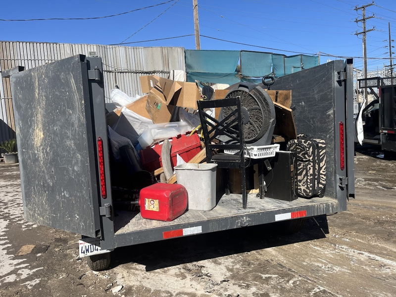 Our dump trailer loaded with a mixed household clear-out on a Los Angeles County job: an office chair, a pedestal fan, a cooler, suitcases, a fuel can and cartons stacked against the headboard