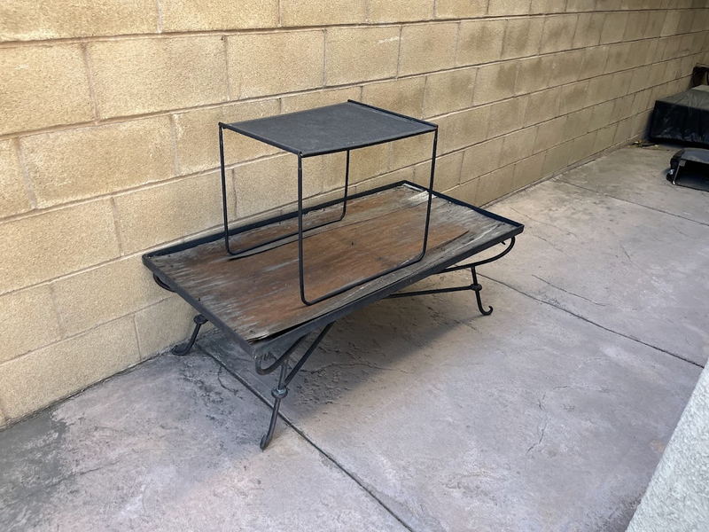 A weathered wrought-iron and timber coffee table and matching side table set out against a block wall on a Los Angeles County patio, waiting to be lifted away