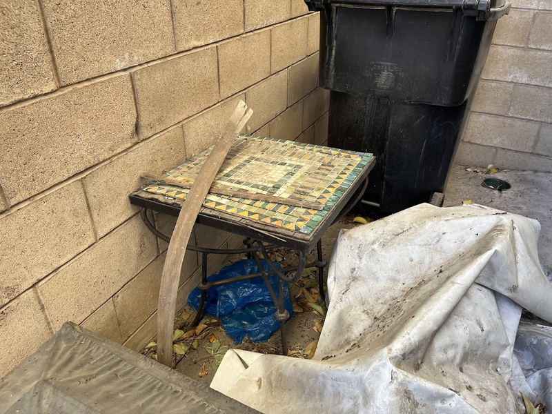 A mosaic-topped patio side table, a torn tarp and bagged waste tucked into the corner of a block-wall side yard beside a wheeled trash bin on a Los Angeles County pickup