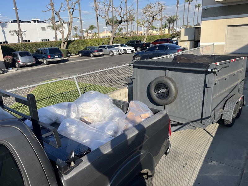Our pickup loaded with bagged waste and hitched to the gray dump trailer, parked against the fence line of a Los Angeles County commercial lot under the palms