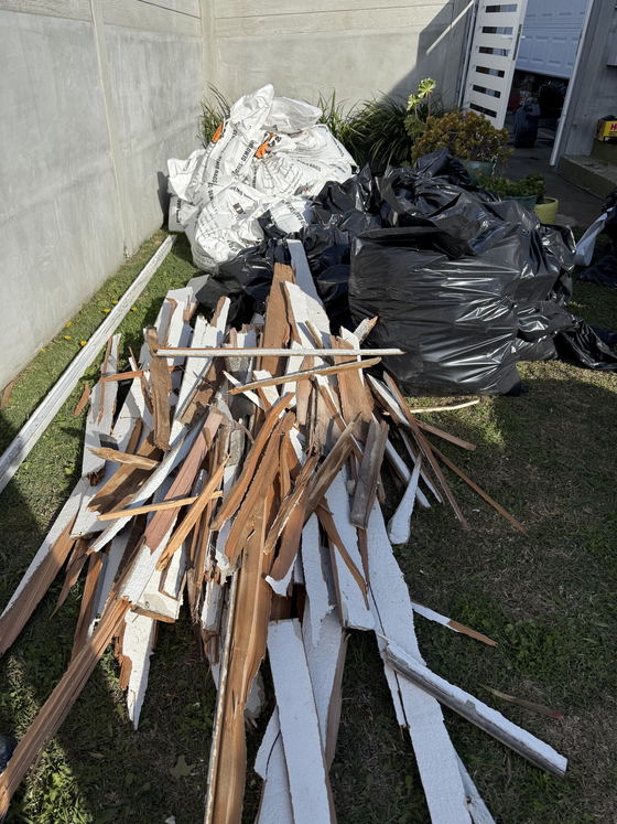 Broken fence boards, splintered lumber and strips of stucco piled on the grass of a Los Angeles County back yard, next to filled white demolition bags and black contractor bags waiting to be carried out